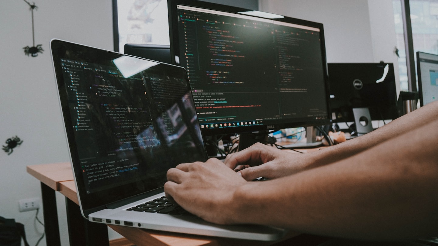 Manos escribiendo código en un portátil conectado a un monitor grande.